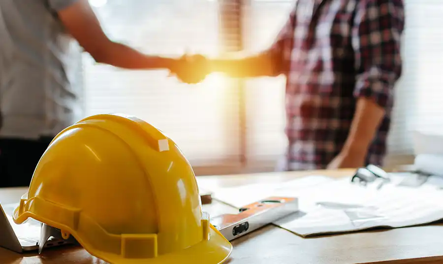 A contractor shaking hands with a client beside a hard hat on the table.