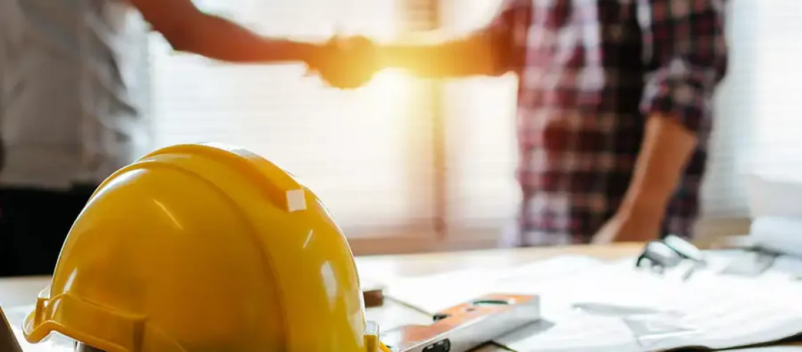 A contractor shaking hands with a client beside a hard hat on the table.
